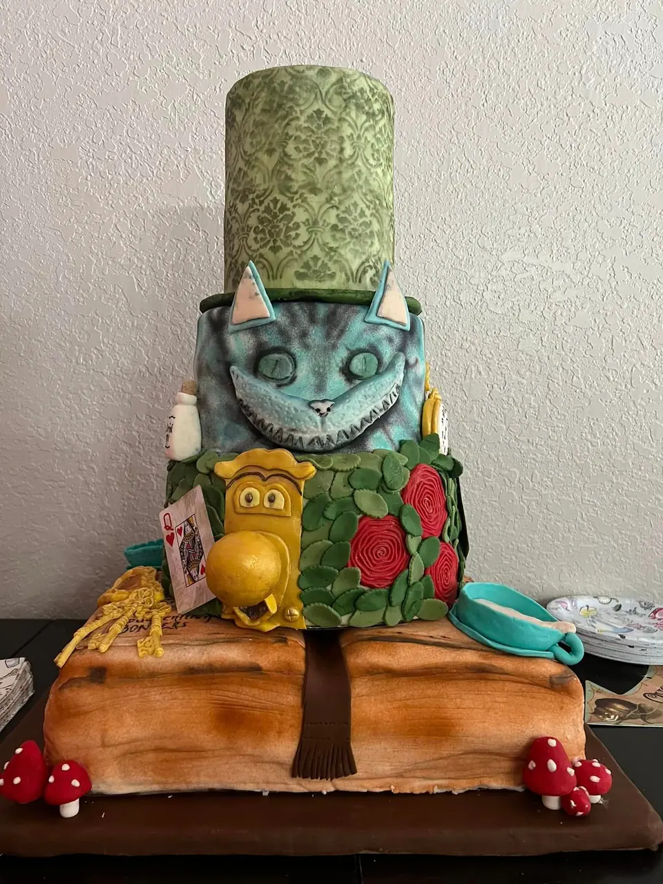 A multi-tiered cake designed for an Alice in Wonderland Party, looking like the Cheshire Cat with a large hat, a pocket watch, and detailed decorations on a wooden-like base.