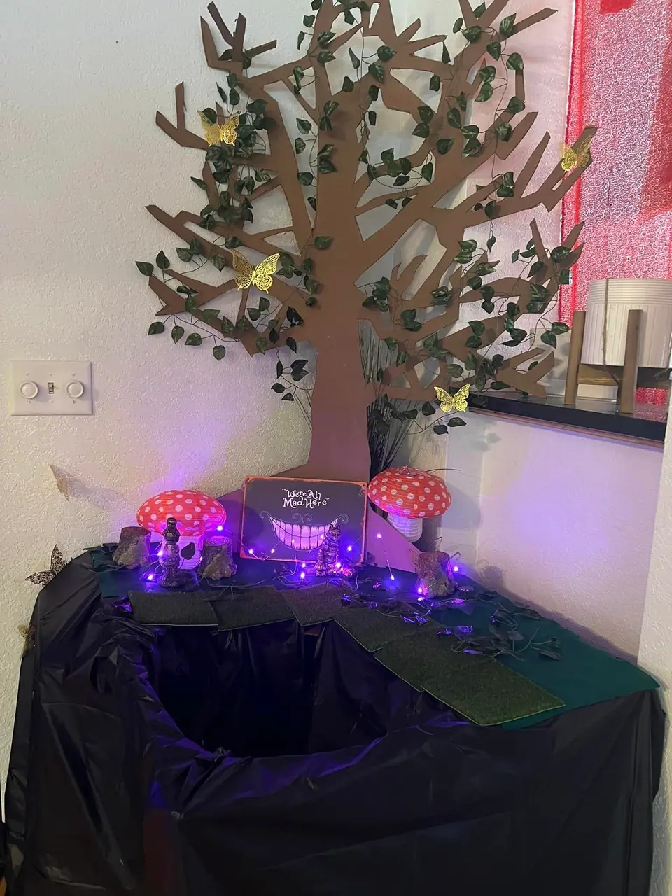 A decorated display straight out of an Alice in Wonderland extravaganza features a handmade tree adorned with paper butterflies, artificial grass, and whimsical mushroom decorations, all illuminated by enchanting purple lights. A "Magic Meadow" sign graces the backdrop.