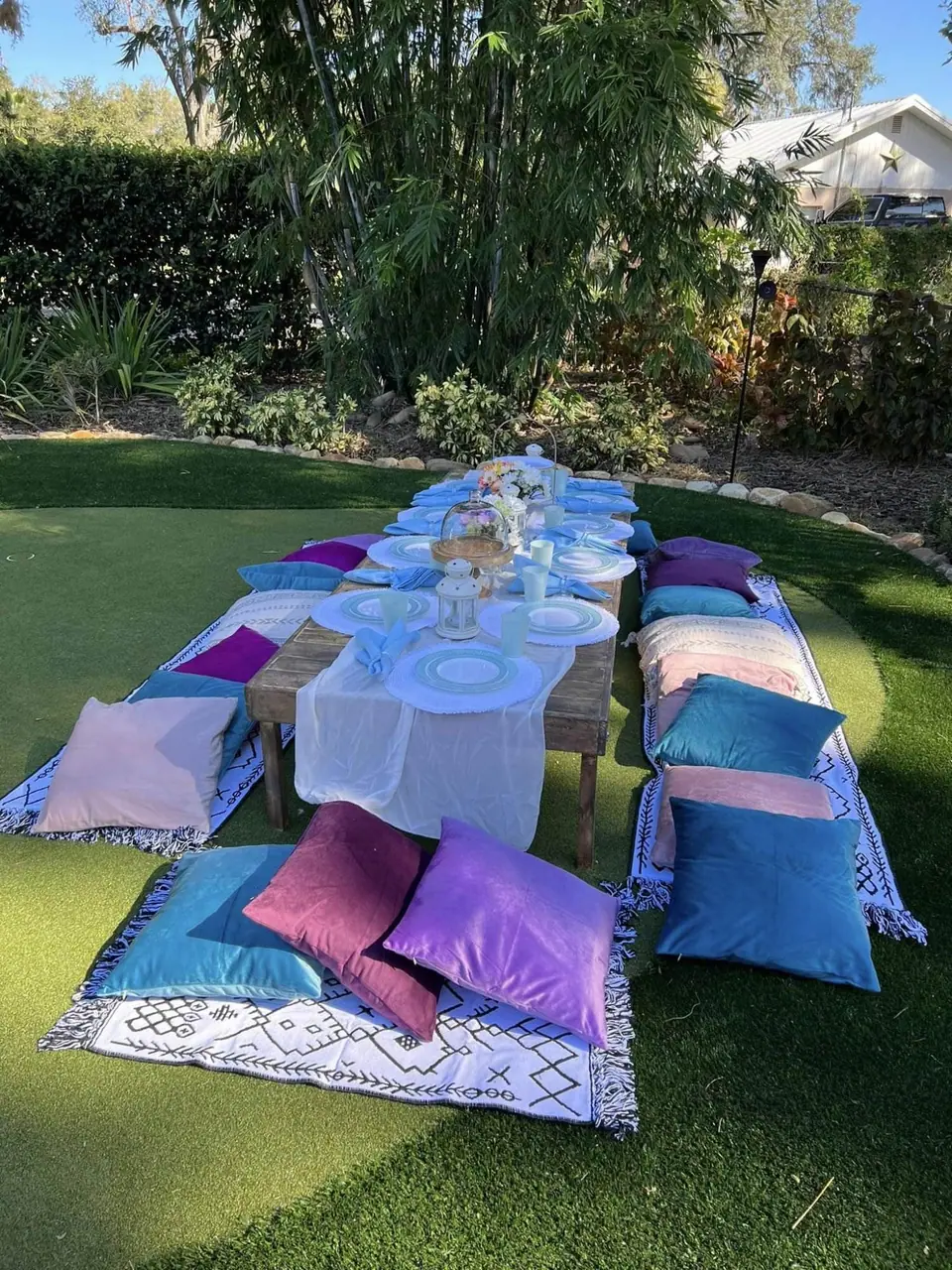 An outdoor garden setup featuring a low wooden table with white plates and floral centerpieces, perfect for a kids party rental. Colorful cushions surround the table, placed on patterned mats on the grass, with a cozy teepee nearby for added charm.