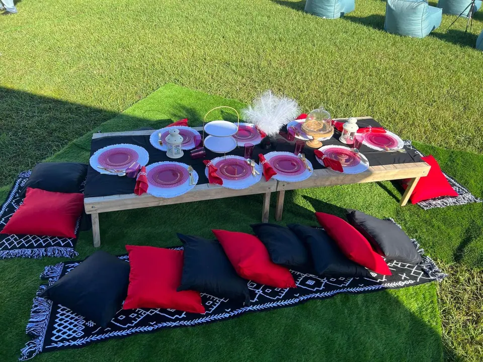 A low wooden table set for a meal on a grassy lawn, perfect for a slumber party. Cushions and mats in red, black, and white surround the table. Plates, drinkware, and covered food items are arranged neatly on the table next to an inviting teepee.