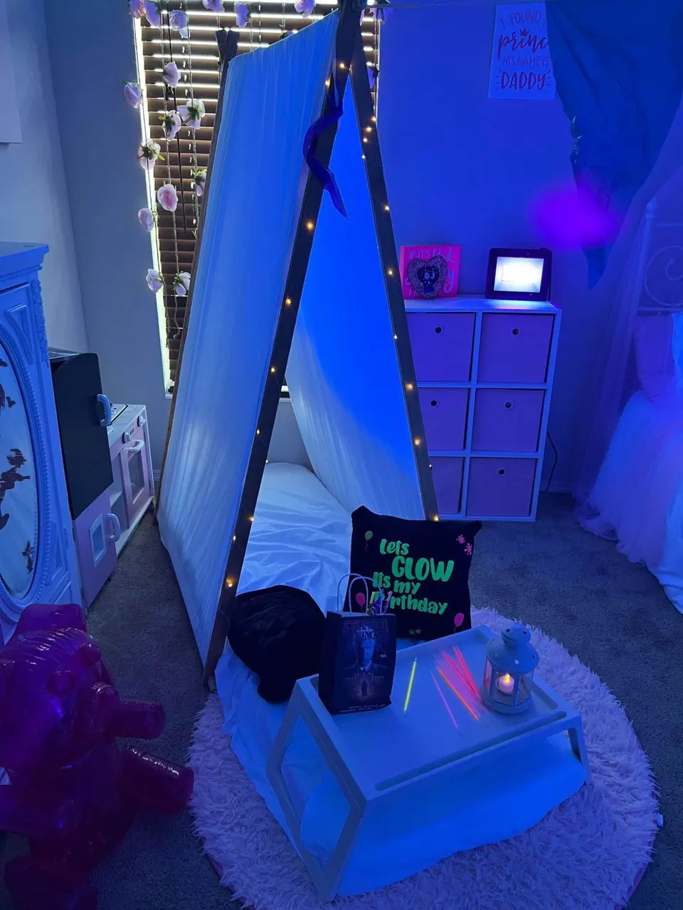 A child's bedroom with a teepee tent illuminated by string lights, surrounded by whimsical decorations and a slumber party-themed setup.