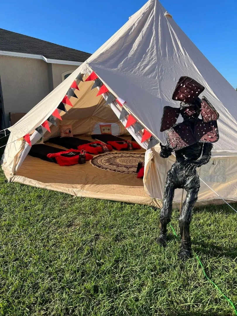Set up the Stranger Things Party Theme: a large canvas sleepover tent with a triangular opening, Demogorgon figure out front, and red bags and pillows inside—ideal for your next Slumberr kids party rental.