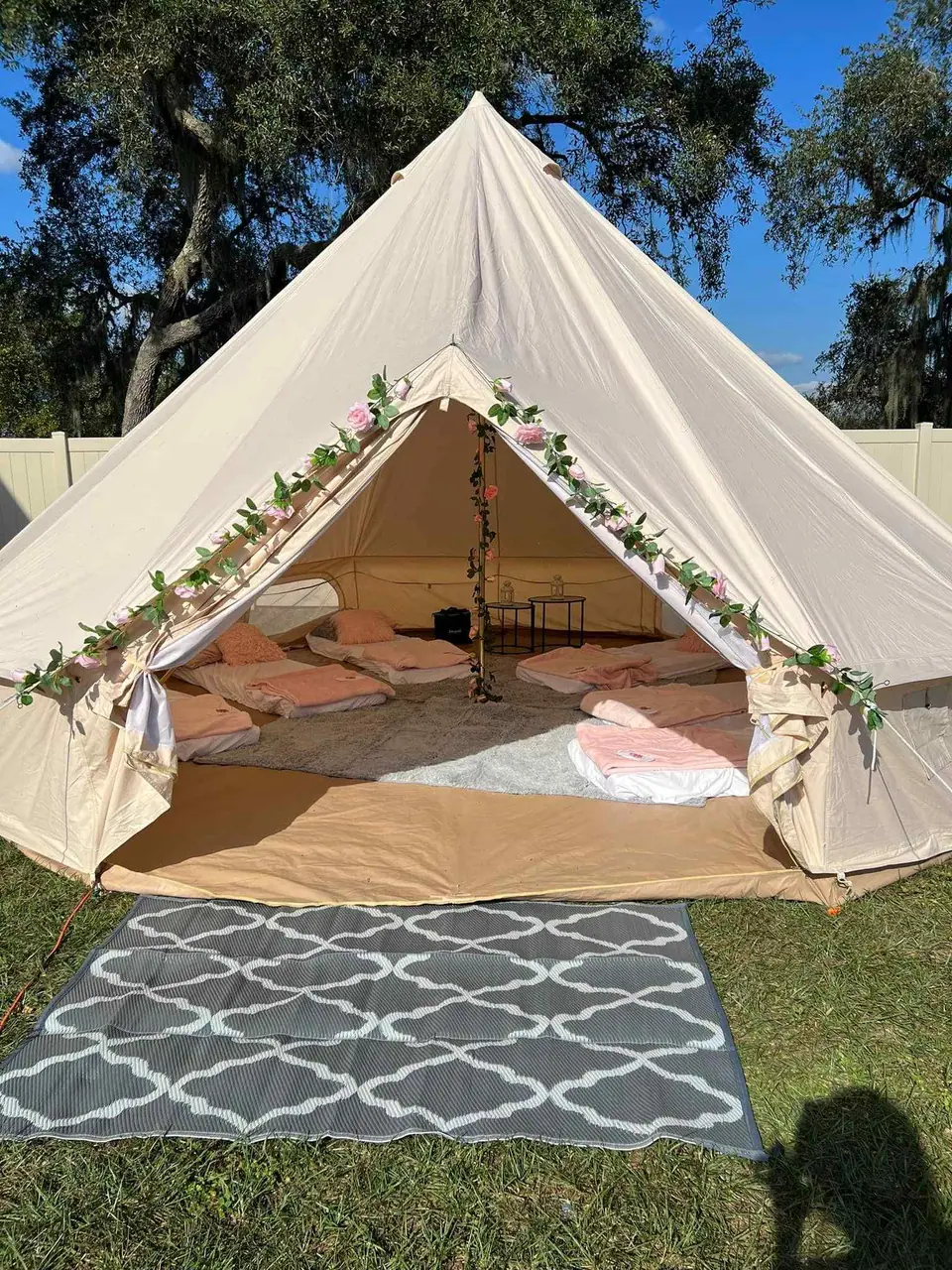 A canvas glamping tent decorated with flowering vines, open to show a cozy interior setup with plush bedding and cushions, set against a backdrop of trees in Central Florida and a clear blue sky.