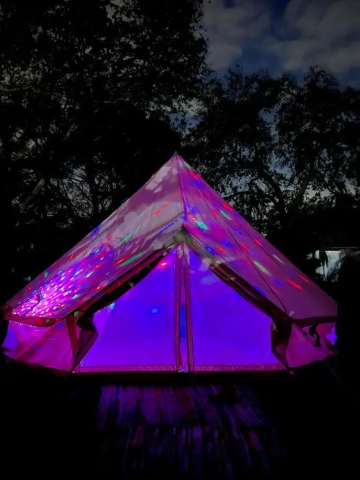 A tent illuminated by vibrant purple and pink lights under a twilight sky hosts a unique slumber party.