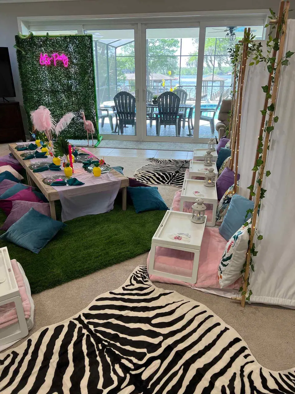A vibrant indoor birthday party setup featuring a grass-like carpet, a long table with assorted colorful cushions, and a pool visible through glass doors.