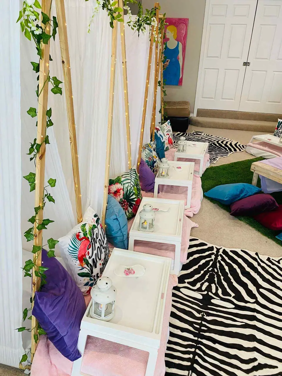Indoor tropical birthday theme picnic setup with colorful cushions, small tables decorated with lanterns, and curtains draped over wooden frames.