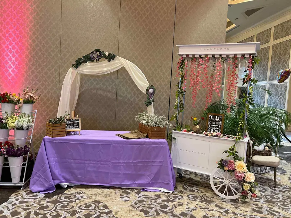 A wedding setup featuring a white arch with floral decorations, a draped table, and a decorative cart with flowers and balloons in an elegant hall for a Central Florida Corporate Event.