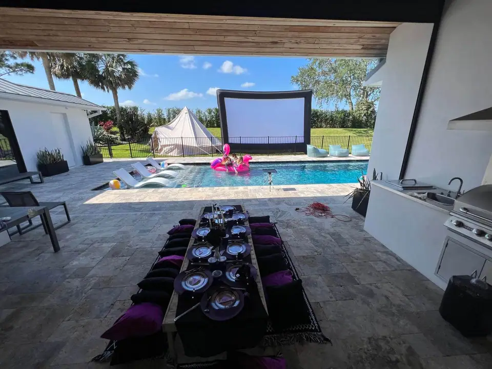 Outdoor pool area with lounge chairs, inflatable flamingo, large movie screen, and a Wednesday Addams Party Theme luxury picnic setup by Slumberr at the dining table under the covered patio.