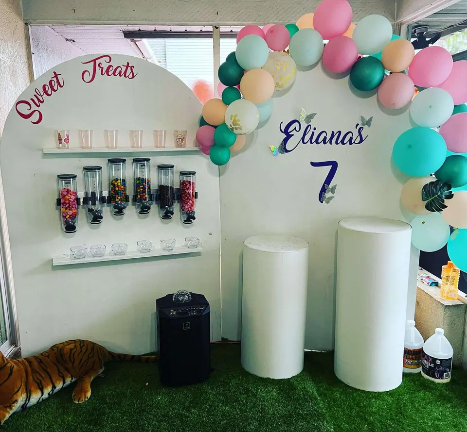 A candy display with clear dispensers, cups, and jars labeled "Sweet Treats" sits next to balloons and a sign reading "Eliana's 7" at an outdoor movie night Florida birthday party.
