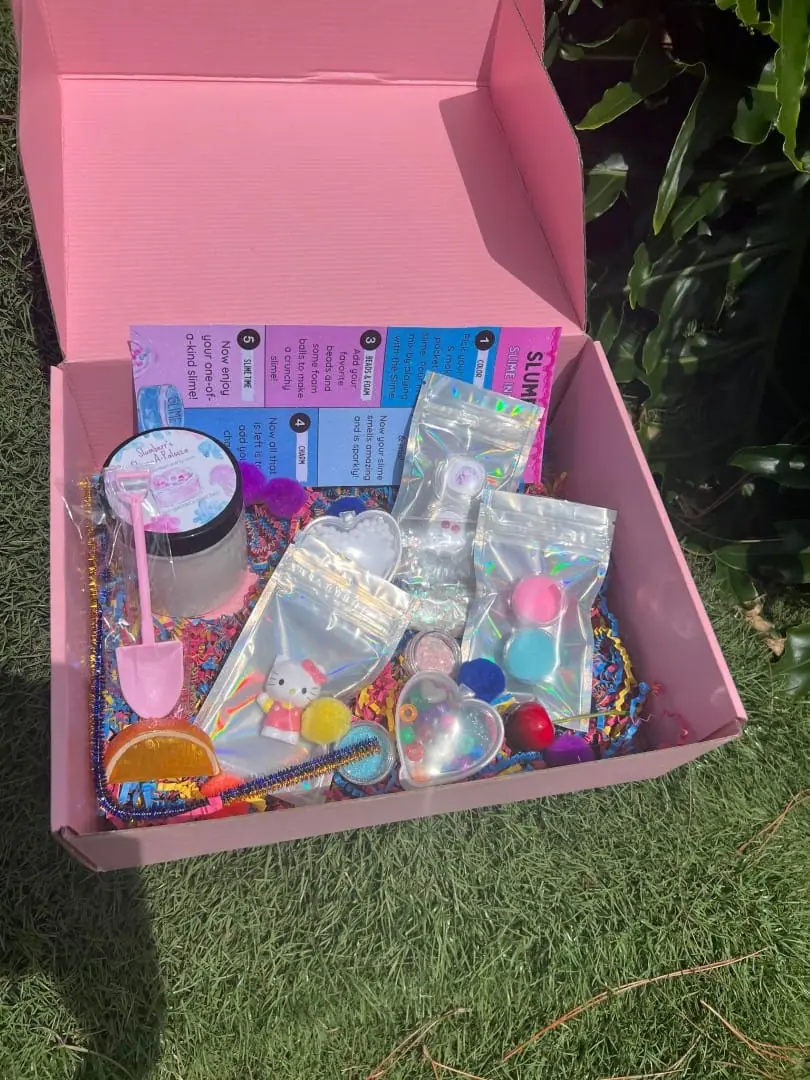 An open pink box containing various colorful children's toys and party favors, including bracelets, a shovel, and small containers, set on grass—delivered to your doorstep.