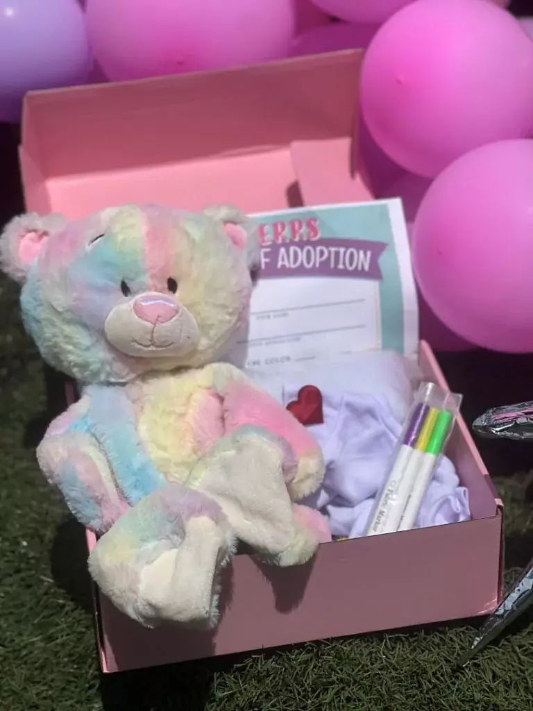 A pastel rainbow teddy bear in a pink box with an adoption certificate, markers, a cloth, and pink balloons in the background—perfect for celebrating special moments. Ideal for St. Petersburg party rentals, this adorable set adds a whimsical touch to any event.
