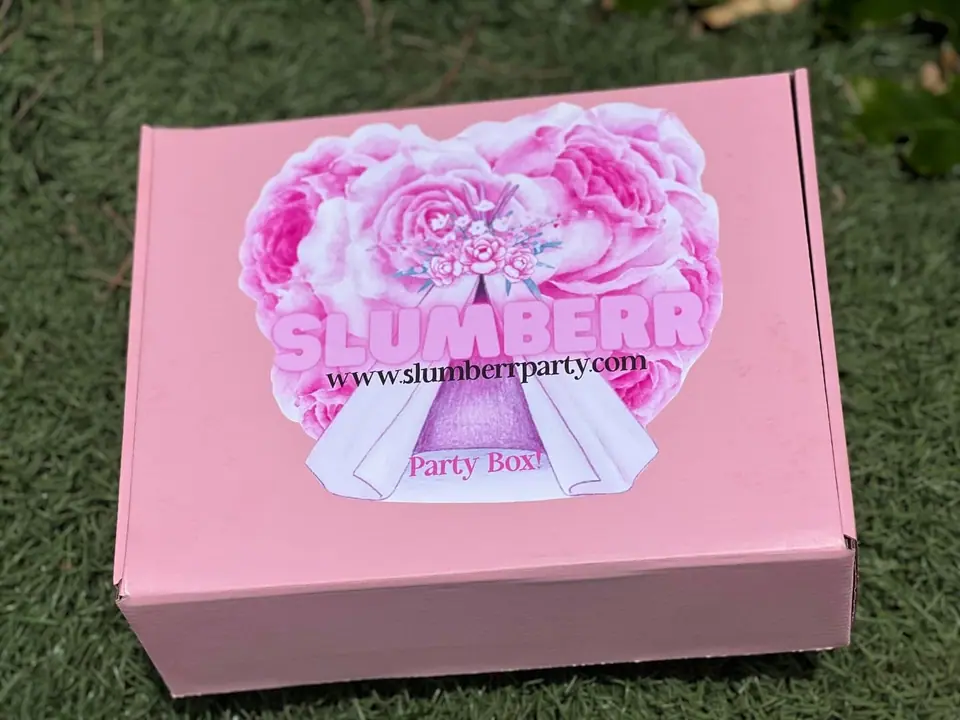 A pink Slumber Party Box with a floral design and the words "Fun Delivered" on top, placed on grass.