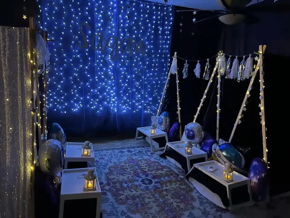 A dimly lit room decorated with a "dream" sign in blue lights, featuring small teepees and celestial-themed cushions on the floor, evoking a magical night.