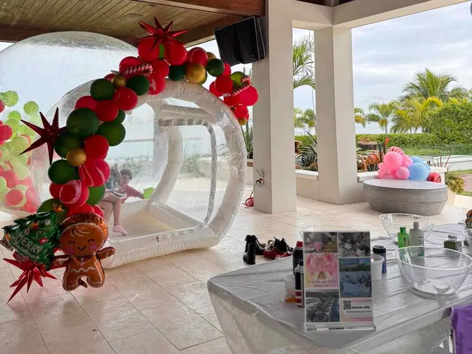 A child sits inside a transparent inflatable dome decorated with balloons, near a table with party supplies from a local party shop and a large outdoor planter with balloon sculptures in South Florida.