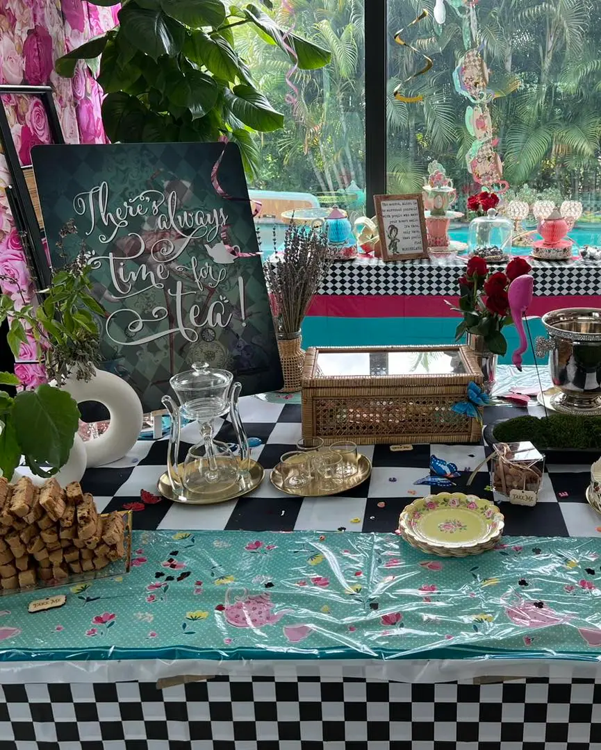 Elegant Alice in Wonderland party setup with a "there's always time for tea" sign, decorated tables with pastries and teapots, and a tropical backdrop.