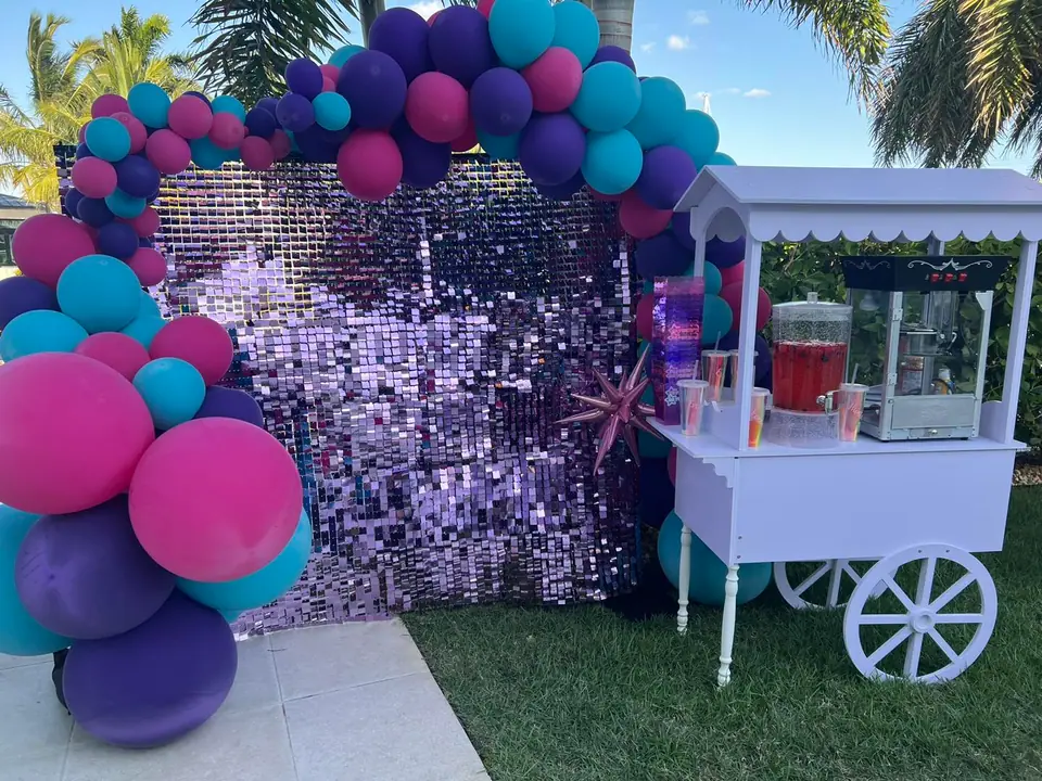 A party setup from a top party shop, featuring a sequined backdrop, colorful balloon arch in pink, purple, and teal, and a white cart with drink dispensers and cups—all beautifully arranged on the grass in Central Florida.