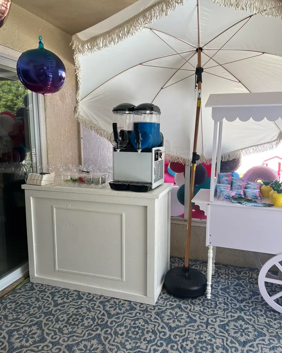 A frozen drink machine sits on a white counter under a large umbrella next to a decorated cart, with plastic cups stacked on the side—perfect for an outdoor movie night in Florida.