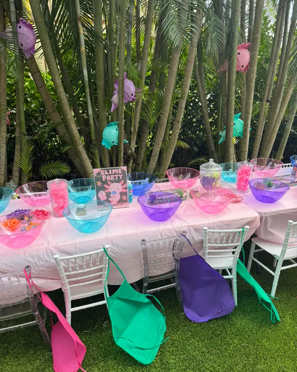 Vibrant outdoor slime party setup featuring colorful bowls filled with an array of slime-making ingredients, neatly arranged aprons draped over chairs, and picturesque palm trees swaying in the background. Perfect for a fun-filled kids' activity day or creative DIY slime workshop, this idyllic setting combines the joy of crafting with the beauty of nature.