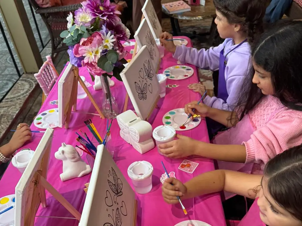 Children sit at a pink-covered table painting butterfly designs on canvases, with paint palettes, brushes, and a vase of flowers in the center—a charming scene perfect for any party planner organizing a creative glamping or teepee-themed celebration.