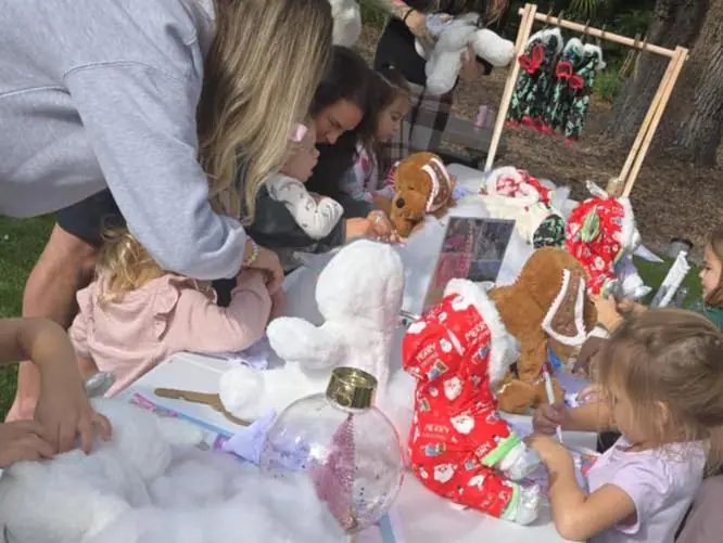 Children and adults gather around a table outdoors, enjoying a fun kids activity as they assemble stuffed animals and decorate them with clothes and accessories.