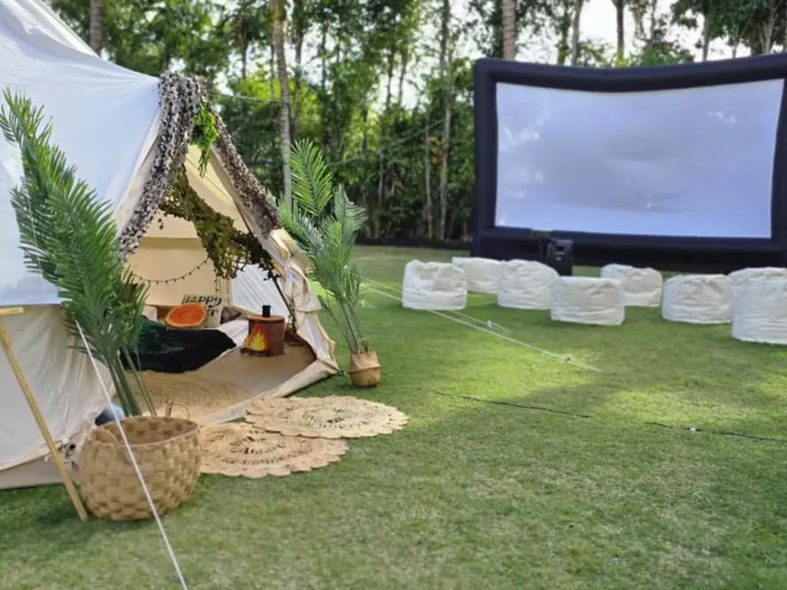 A white tent with cushions and decorative plants is set up on grass for an outdoor movie night Florida style, next to an inflatable screen with round seats arranged in front.