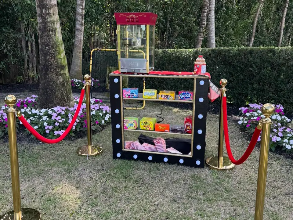 A small concession stand with a popcorn machine, boxed candies, and popcorn bags is set up for an outdoor movie night in Florida, bordered by red rope barriers and surrounded by flowers and lush greenery.