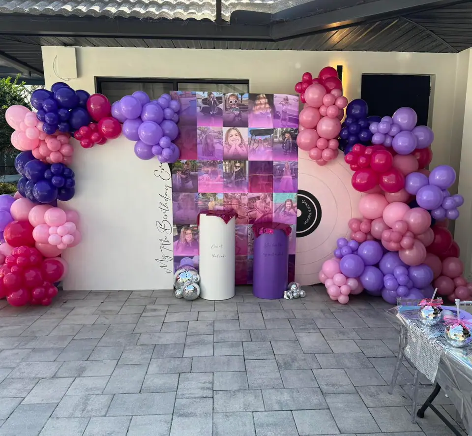 Balloon arch in shades of pink, purple, and red surrounds a photo backdrop and two decorative columns at an outdoor celebration event with a tiled floor—perfect for any Central Florida party shop setup.