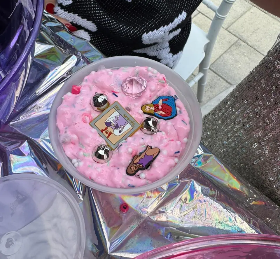 A round container filled with pink, textured slime—perfect for a kids activity—is decorated with metallic beads, a large gem, and three cartoon pins, sitting on a shiny, iridescent surface.