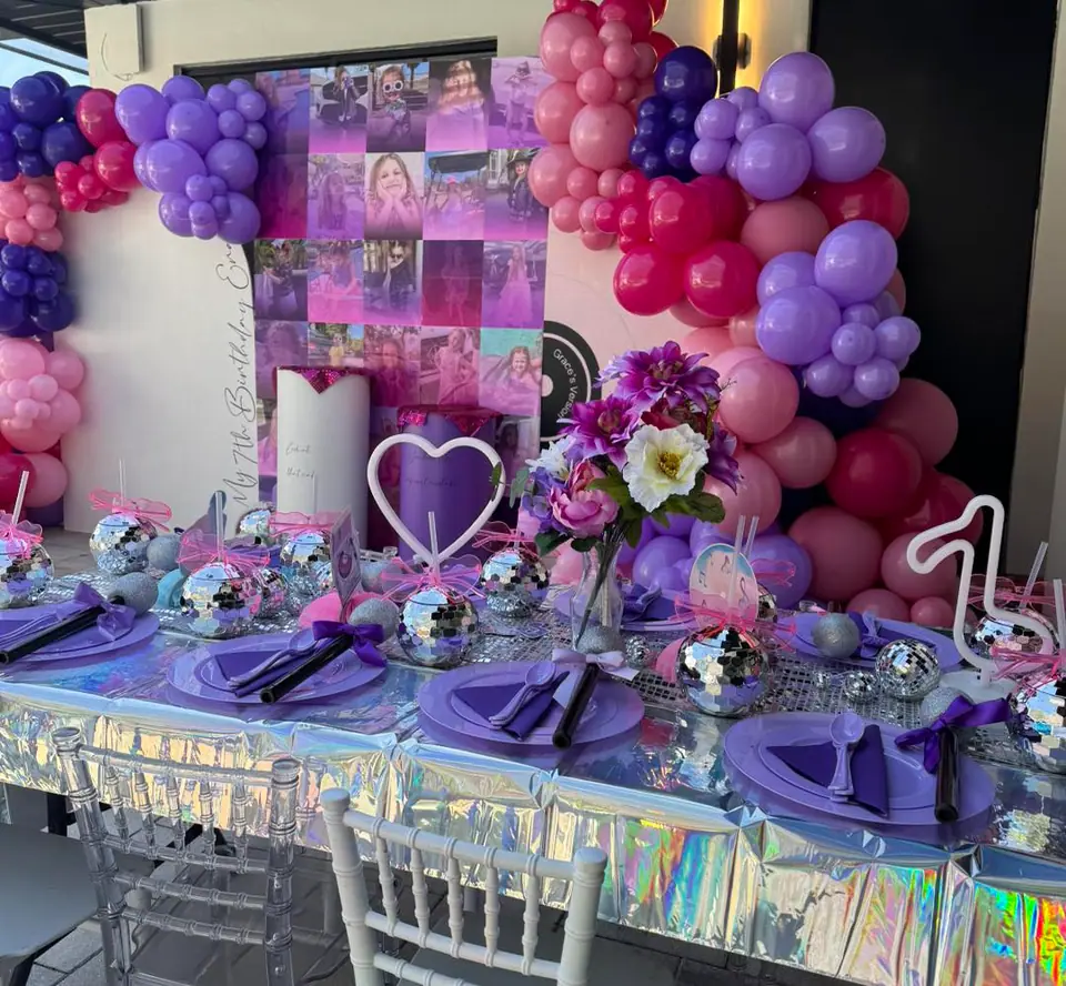 A party table with purple plates, disco ball centerpieces, and iridescent tablecloth sets a festive mood for an outdoor movie night Florida, complete with pink and purple balloons and a collage backdrop decorated with photos and flowers.