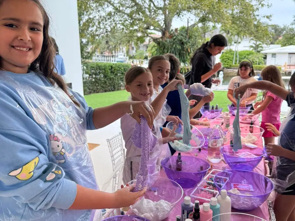 A group of children gather around a table at a party shop, making slime with purple bowls, craft materials, and bottles, while smiling and showing their creations.
