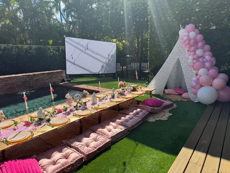 Outdoor garden setup by the pool with low picnic tables, pink cushions, a white teepee adorned with pink balloons, and a large projector screen—perfect for your next celebration from a top party shop in South Florida or Central Florida.
