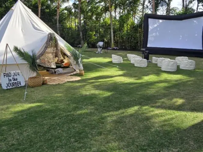 Outdoor movie party setup with a tent, a sign saying "Find Joy in the Journey," white seats arranged on grass, and a large inflatable movie screen in front of trees.