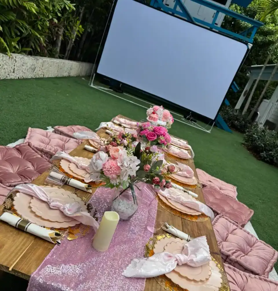 A picnic-style table set with pink cushions, floral centerpieces, and a sequin runner, arranged outdoors on grass in front of a large blank projector screen—perfect for gatherings in Central Florida or styled by your favorite party shop.