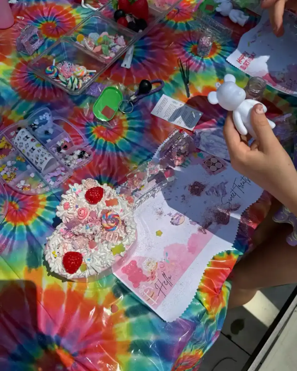 Hands holding a white bear figurine over a tie-dye table filled with craft supplies, decorated pastries, and art materials—a perfect kids activity for creative fun.