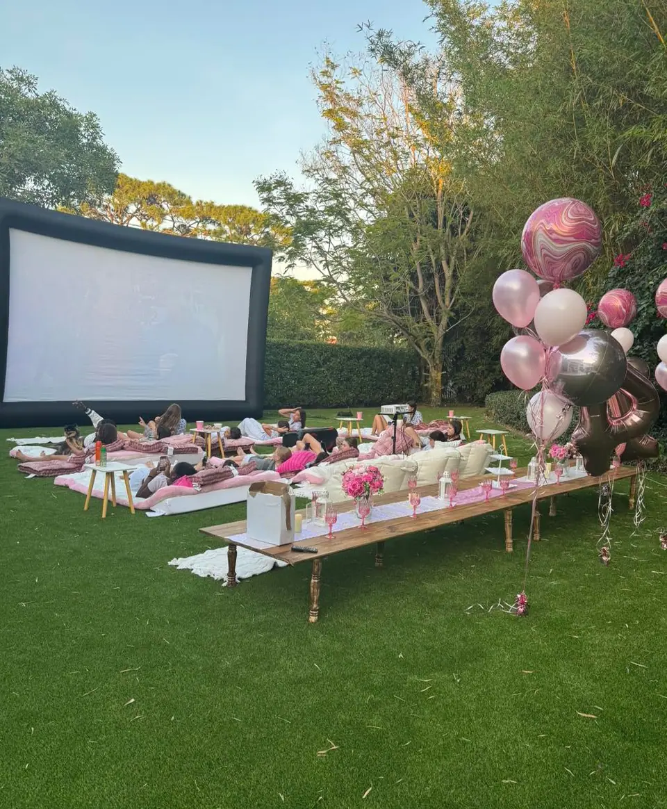 Outdoor movie night setup with people lying on cushions, a large screen, low tables with flowers, and pink and silver balloons on a grassy lawn creates the perfect outdoor movie party atmosphere.