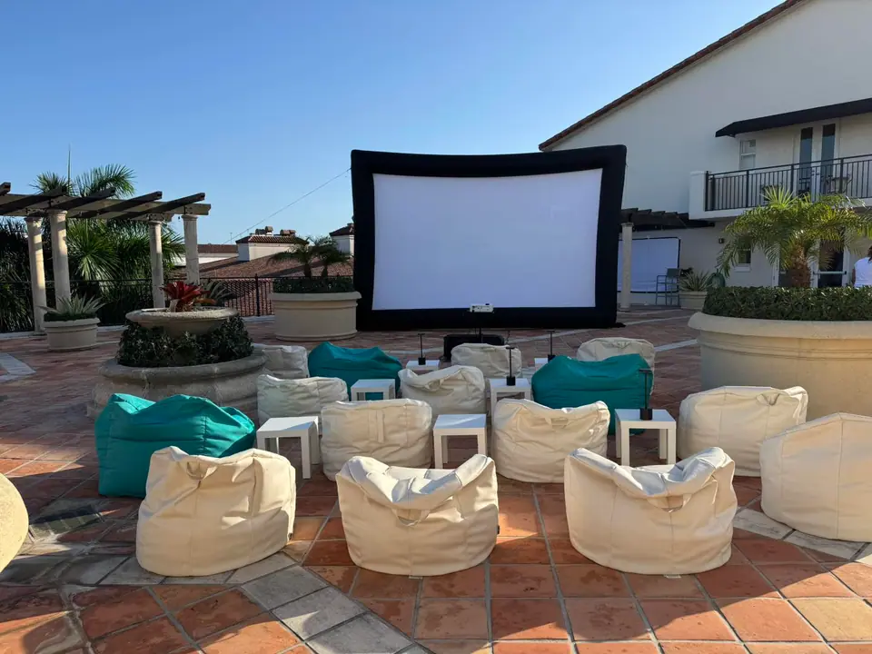 An outdoor movie party setup with a large screen, projector, and rows of white and teal bean bags arranged on a tiled patio under a clear sky.