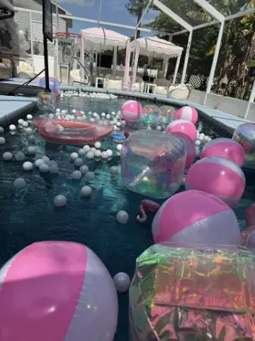 A swimming pool filled with pink and white beach balls and inflatable floats sets the perfect scene for a beach cabana party, with stylish white outdoor cabanas and comfortable seating in the background.