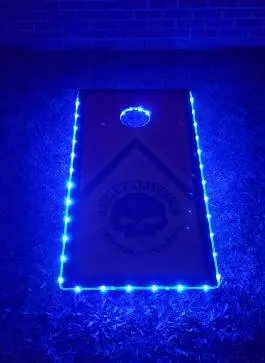 A cornhole board lit with blue LED lights, featuring a design with a skull and text, perfect for a lively kids party rental.