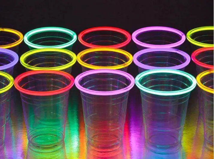 A row of transparent plastic cups with neon-colored rims glow under blacklight on a reflective surface, setting the perfect vibe for a slime party.
