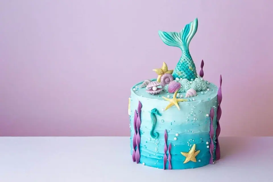 A mermaid slumber party-themed cake with a blue icing base, decorated with seashells, starfish, and a teal mermaid tail on top against a light pink background.