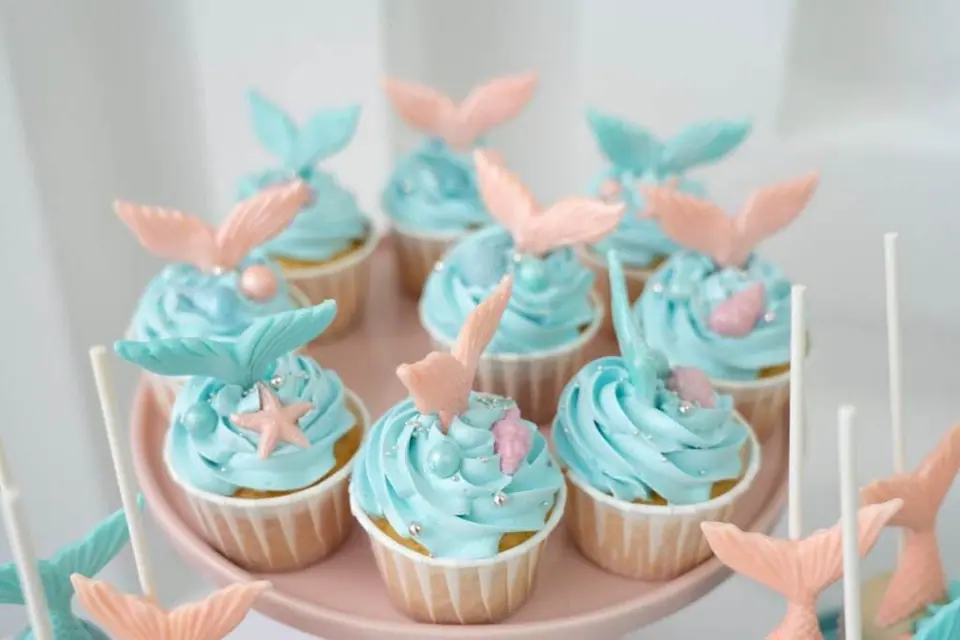 A tray of cupcakes decorated with blue frosting and topped with pink and blue fondant seashells and starfish, perfect for a Mermaid Slumber Party, on a pink background.