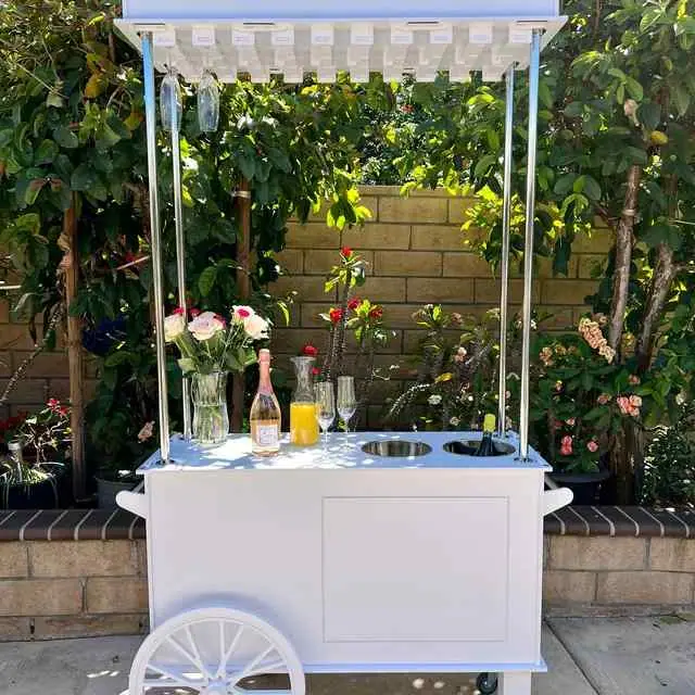 A white cart with a canopy holds a vase of flowers, champagne, orange juice, and glasses—perfect for a glamorous Mother’s Day lounge party in a garden with lush foliage and a brick wall.