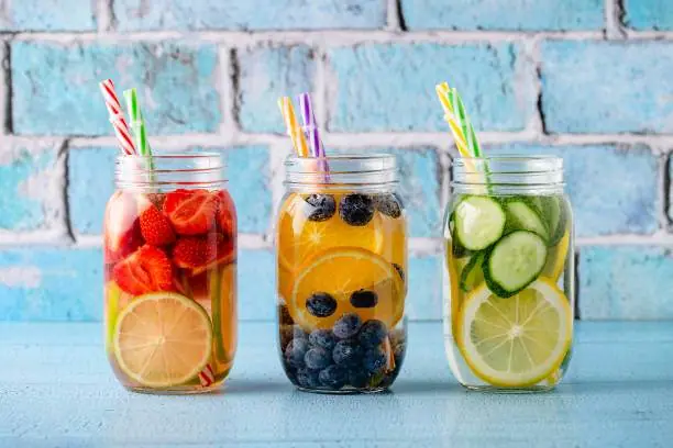Three mason jars filled with colorful fruit-infused water: strawberries and lemon, blueberries and orange, cucumber and lemon. Each jar has patterned straws. Perfect addition to any party rental setup against a blue brick background.