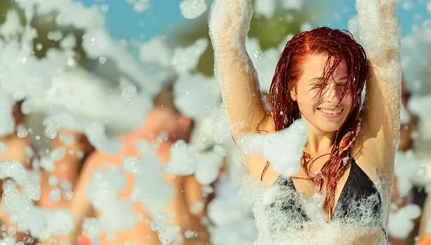 Woman smiling in a black bikini top, enjoying a foam party outdoors, with party rental equipment and other people covered in foam in the background.