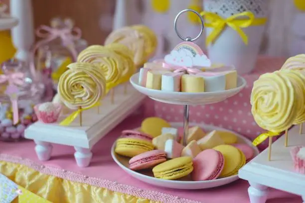 A dessert table featuring pink and yellow macarons, white and yellow swirl meringues on stands, and various candies in jars decorated with ribbons, perfect for a kids party rental.
