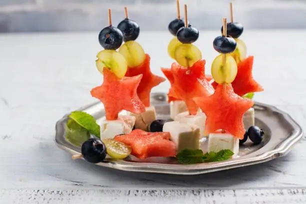 Skewers with star-shaped watermelon slices, green grapes, and blueberries are beautifully arranged on a plate with mint leaves and cheese cubes—perfect for a kids' party rental.