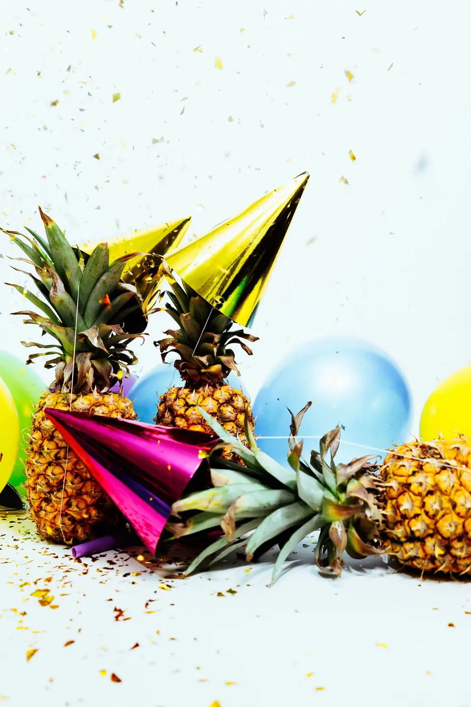 Two pineapples wearing party hats surrounded by colorful balloons and confetti on a white background, celebrating a tropical birthday party.