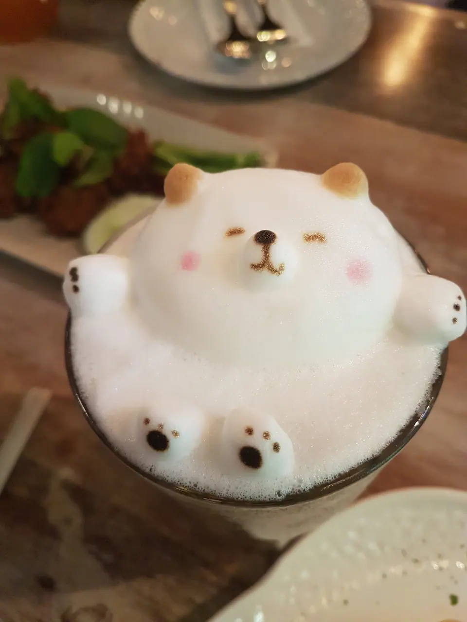 A latte features 3D latte art depicting a bear with eyes closed and arms extended, as if it's just waking up from a magical slumber party. In the background, a dish with food and utensils on a plate is visible.
