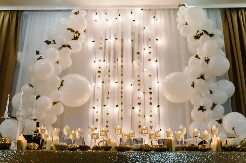 Elevate your baby shower experience with the ultimate event rentals that feature a stunning decorated backdrop. This captivating display boasts oversized white balloons, flowing translucent curtains embellished with twinkling fairy lights, and shimmering gold stars. Set against this enchanting scene is a luxurious table adorned in a glittering gold tablecloth, showcasing an array of beautifully lit candles nestled in elegant glass holders. The sophisticated arrangement creates an unforgettable and elegant atmosphere perfect for any memorable celebration. Ideal for those looking to enhance their baby shower decor with elegance and charm.