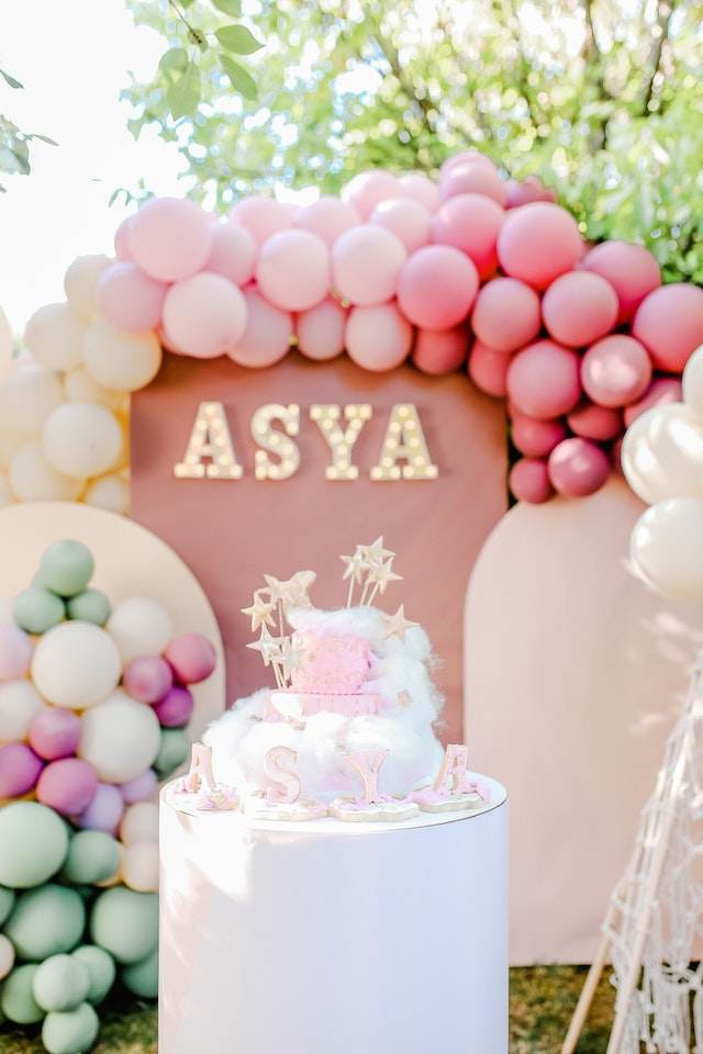 A cake on a pedestal with the name "ASYA" in front, surrounded by pink, white, purple, and green balloons. The background features a decorative arch with the name "ASYA" in lights—an ideal setup for a magical kids party rental or even an enchanting slime party.
